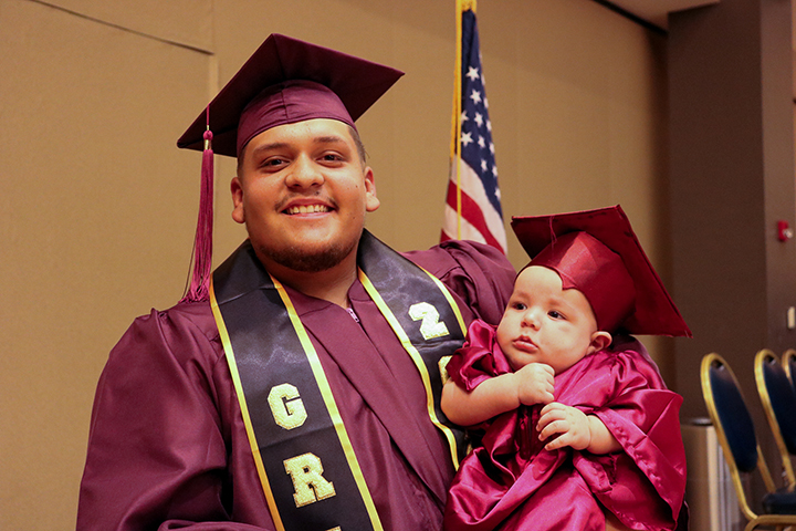 A graduate proudly holds his infant, also wearing a graduation cap and gown