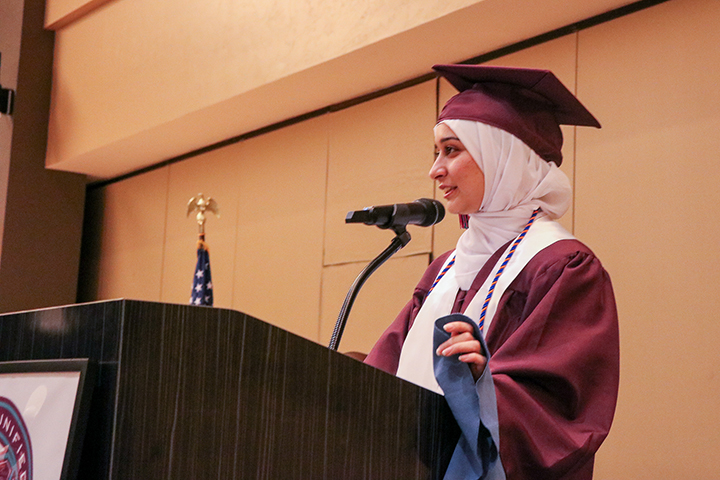A teen girl in a head covering and cap and gown addresses her fellow graduates