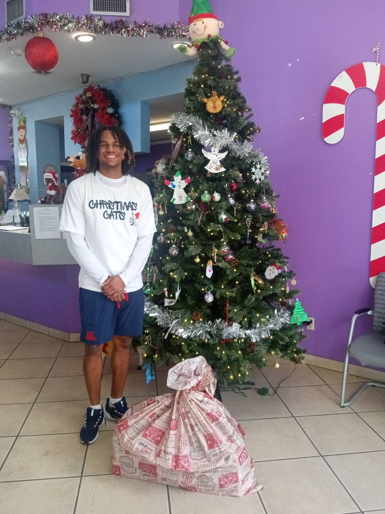 A young man in a Christmas Cats shirt smiles in front of a Christmas tree with a bag of gifts at his feet